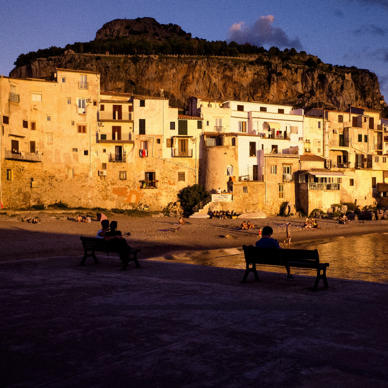 Panoramic sunset view of traditional white buildings nestled against rocky cliffs in a Mediterranean coastal town.