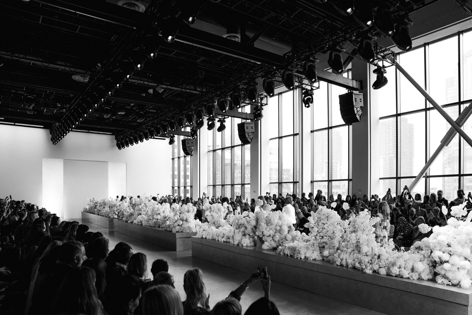 Black and white photo of a fashion show runway lined with white flowers and seated audience.