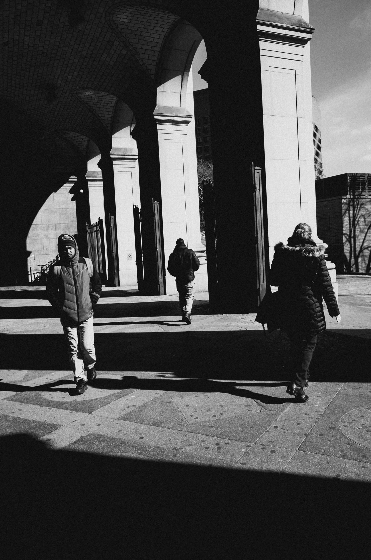 Black and white architectural series showing people walking through a colonnade casting dramatic shadows on the ground.