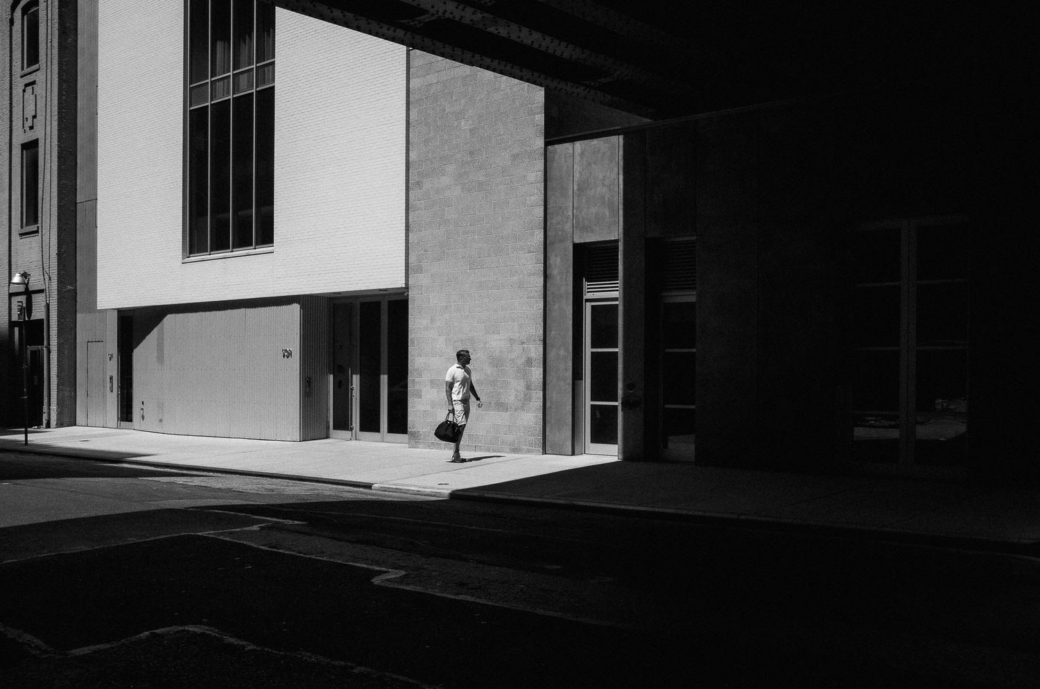 A black and white architectural photograph shows a figure walking past a modernist building entrance in stark sunlight.