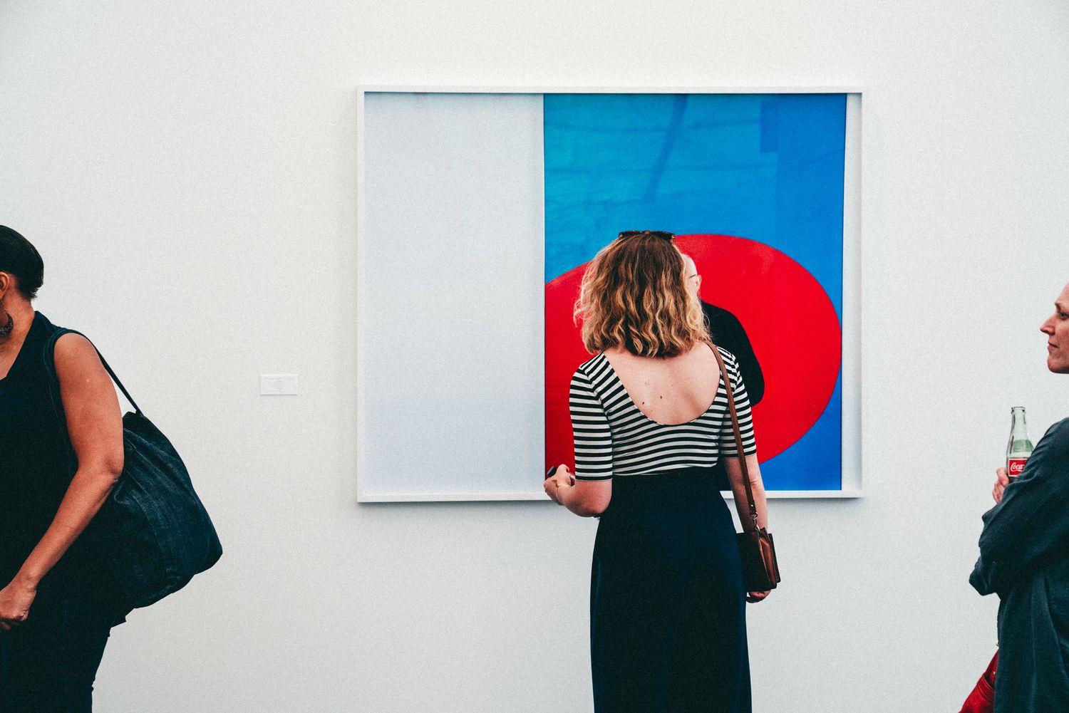Gallery visitors viewing a large colorful abstract painting with red circle and blue shapes on white wall.