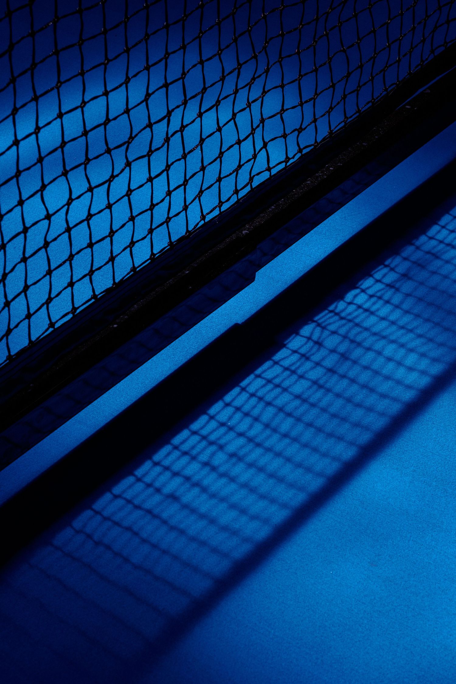 Tennis net silhouetted against blue court surface creating dramatic shadows and patterns.
