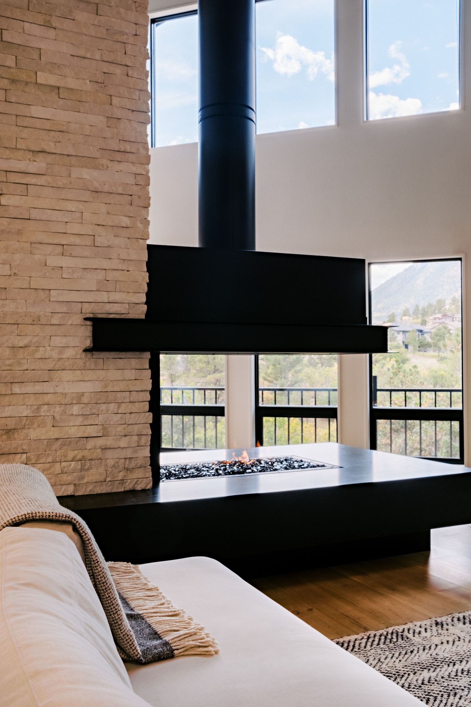 Minimalist living room with stone accent wall and black suspended fireplace overlooking mountain landscape.