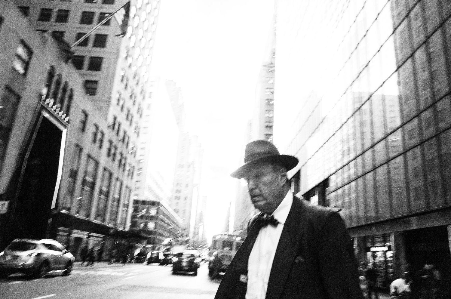 Black and white street photograph of a person in a fedora and bow tie walking past tall buildings in an urban setting.