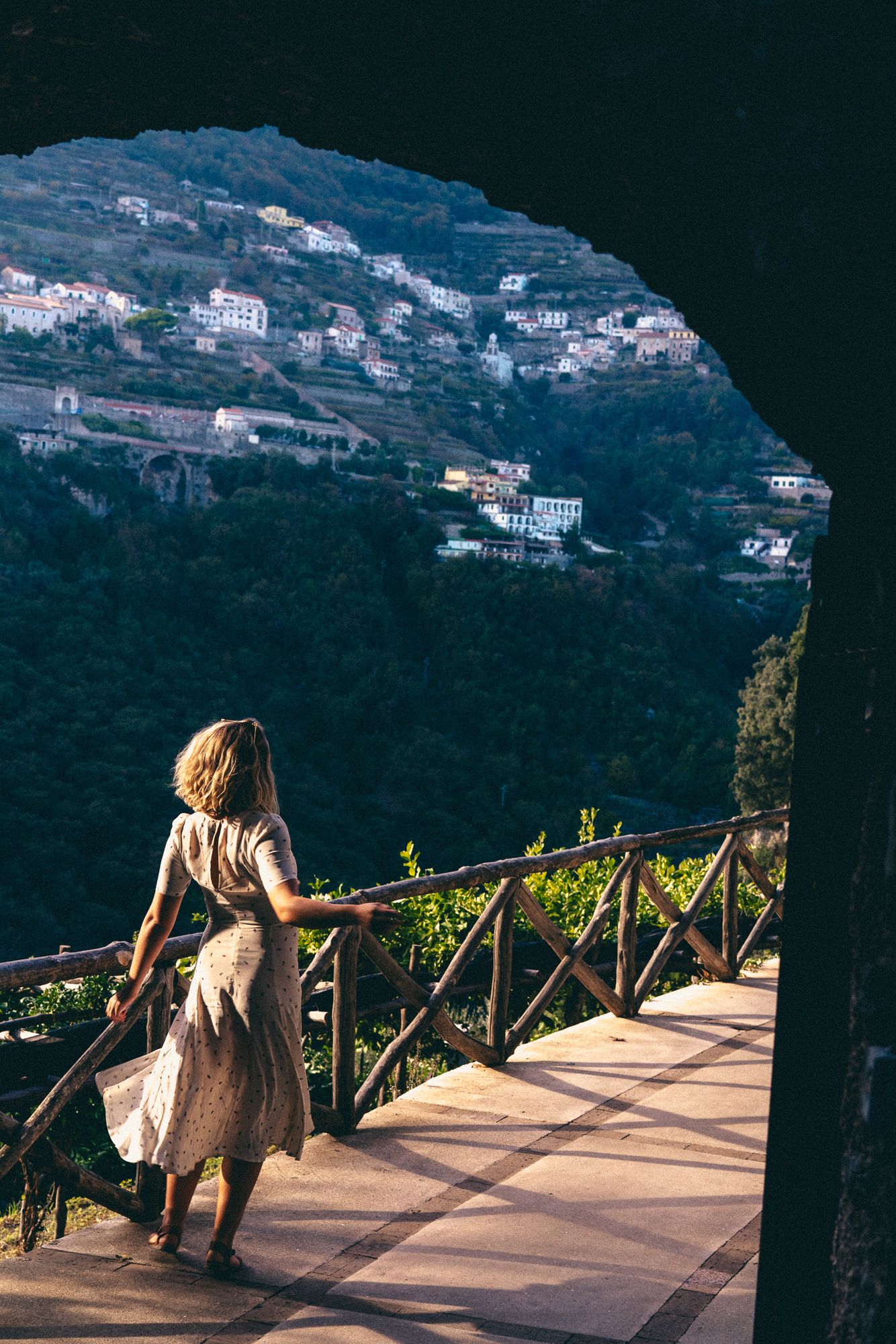 A person in a flowy dress stands on a scenic terrace overlooking the hillside villas of Ravello, Italy at sunset.