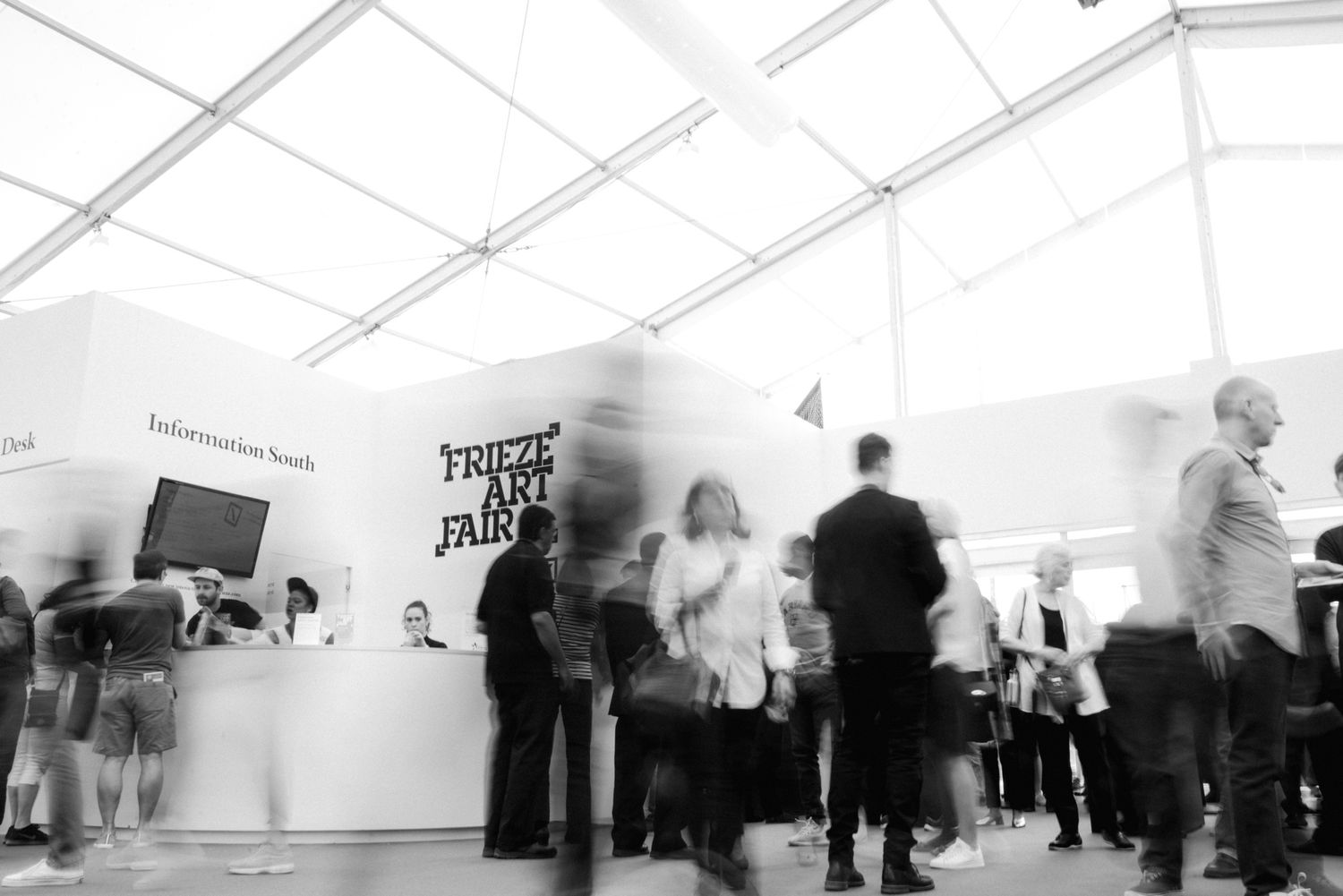 Blurred black and white photo of visitors at the Frieze Art Fair entrance booth under a white tent.