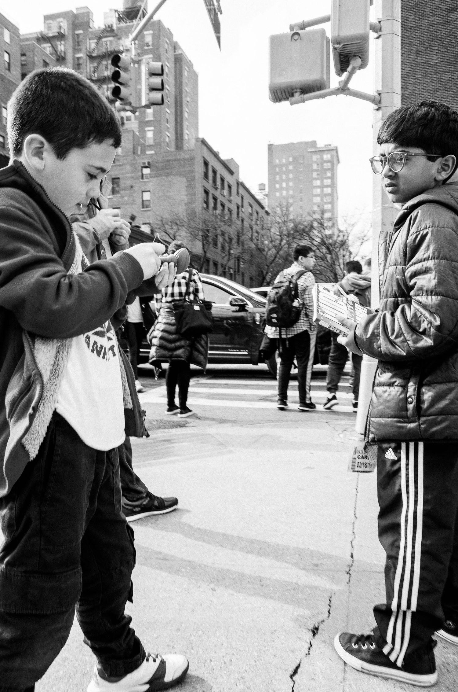 Two children in athletic wear interact on a city street corner with tall apartment buildings in the background.
