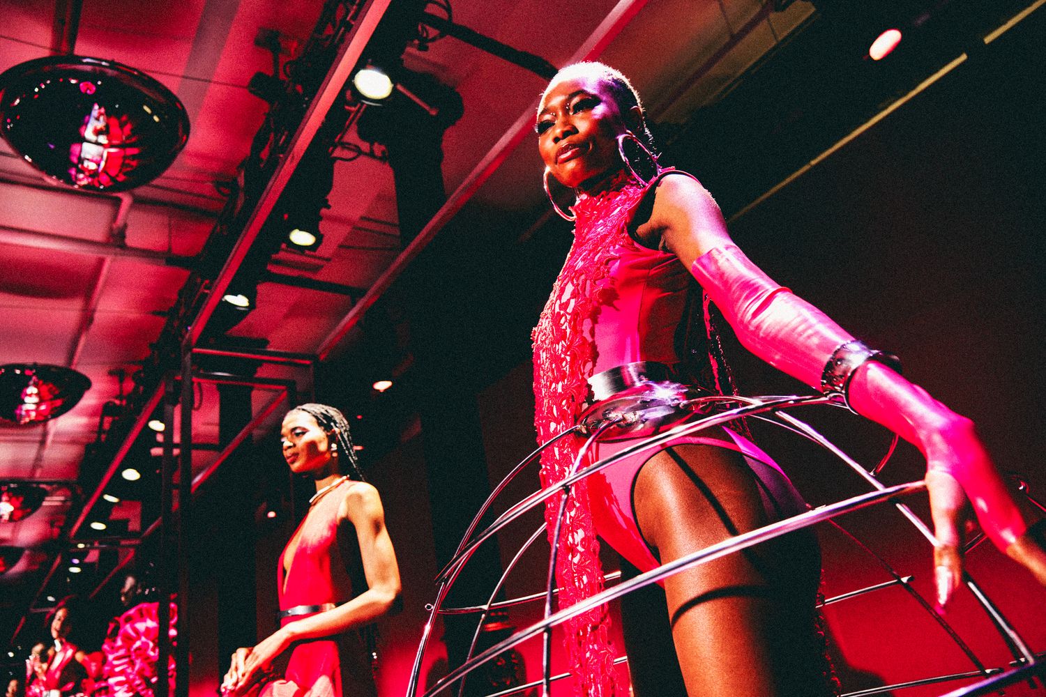 Models in red sequin outfits perform with hoop props under dramatic red lighting at a fashion show event.
