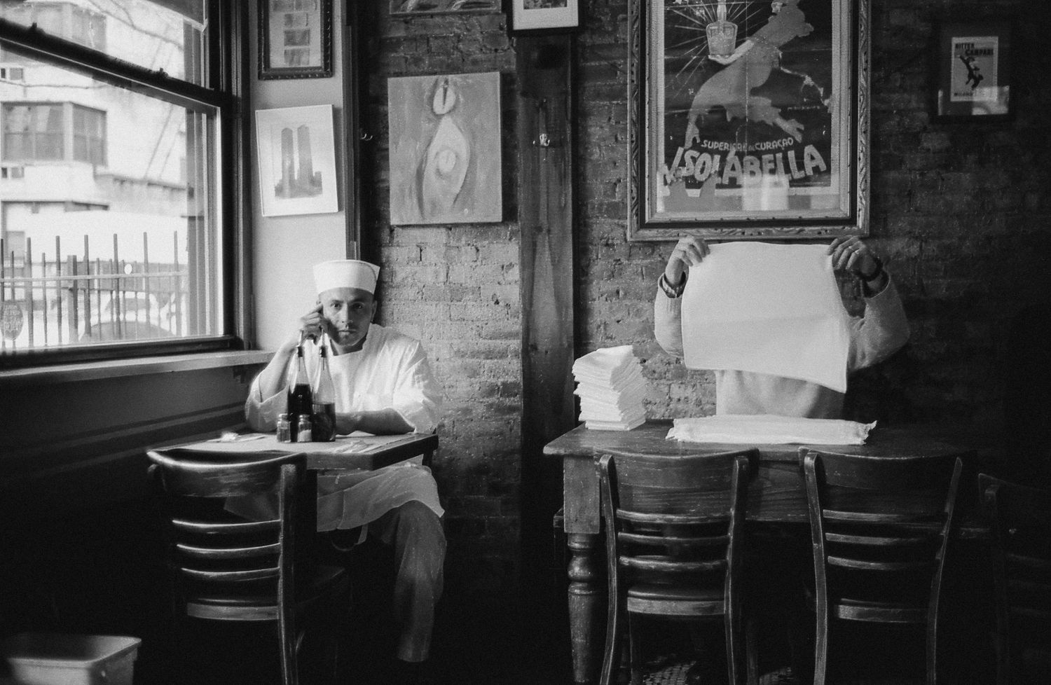 Black and white photo series of empty chairs and tables in a dimly lit cafe interior with framed artwork on brick walls.
