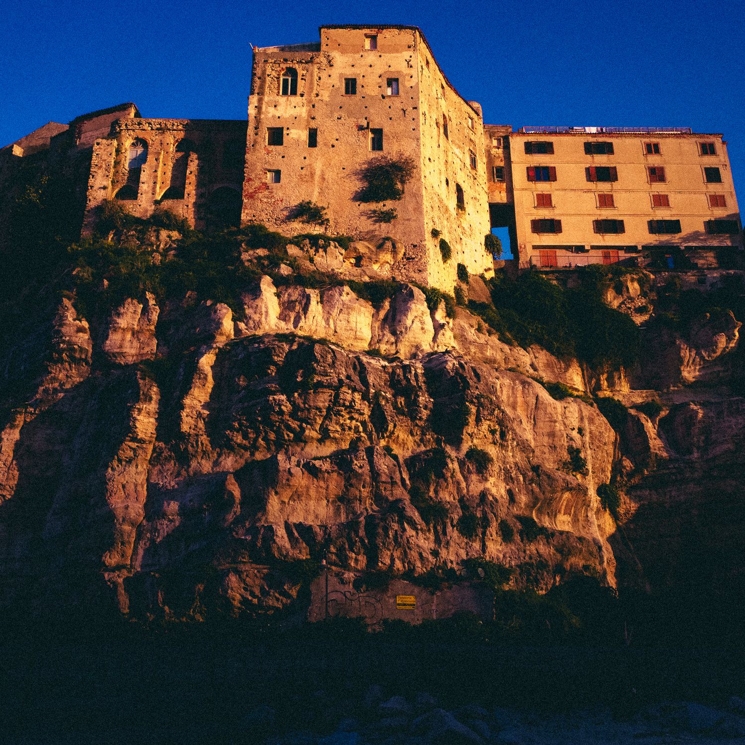 Ancient stone buildings perched dramatically on a steep cliff edge glow golden in warm sunlight against deep blue sky.