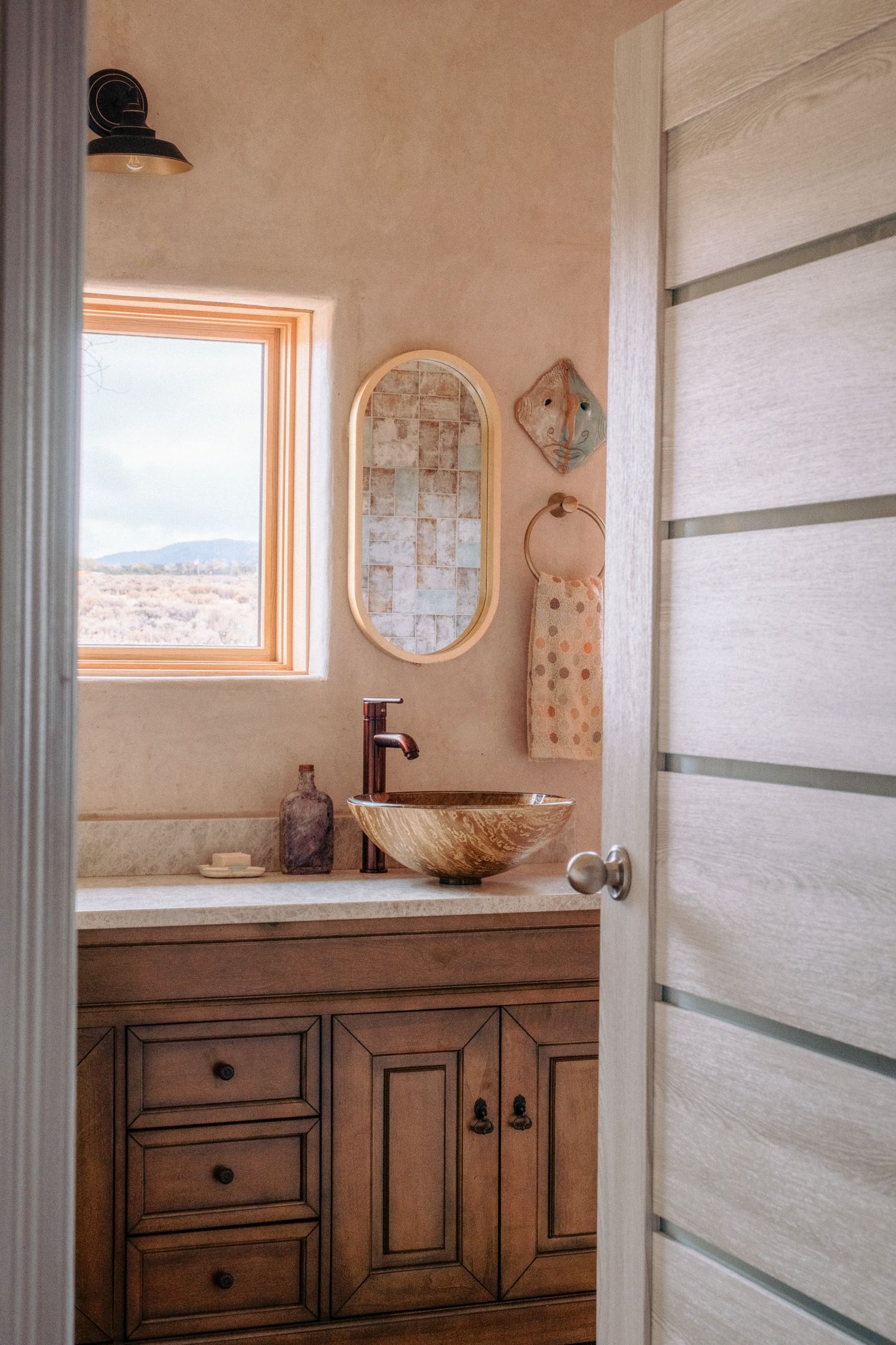 Modern bathroom interior with vessel sink, wooden vanity and oval mirror near window with desert view.