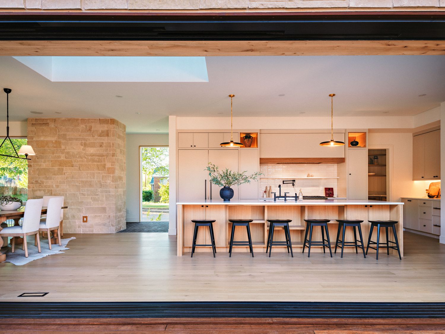 Modern open-plan kitchen with large island, bar stools, pendant lights, skylight, and stone accent wall with seamless indoor-outdoor flow.