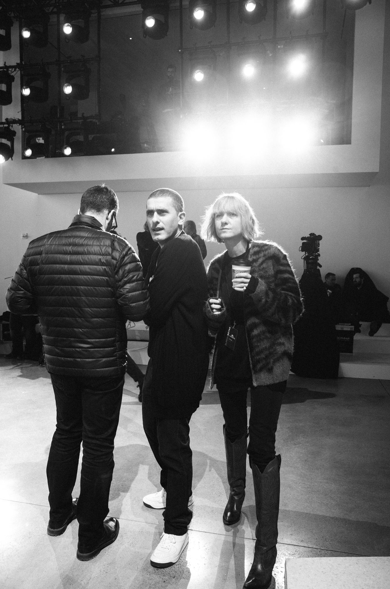 Black and white photo of three people standing indoors under bright stage lights at what appears to be a fashion event.