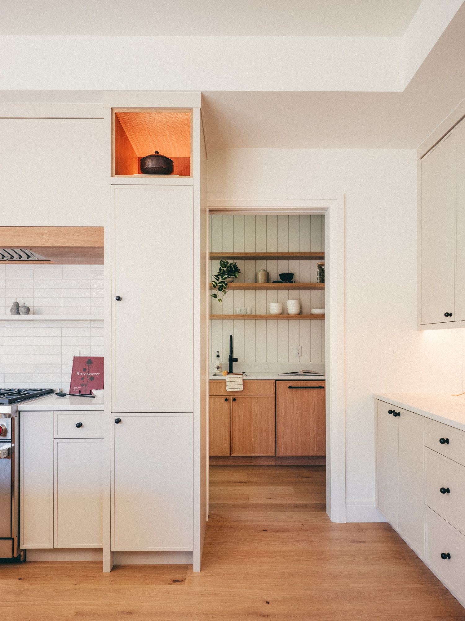 Modern kitchen with white cabinets, wood shelving, black hardware, and a butler's pantry visible through an archway.