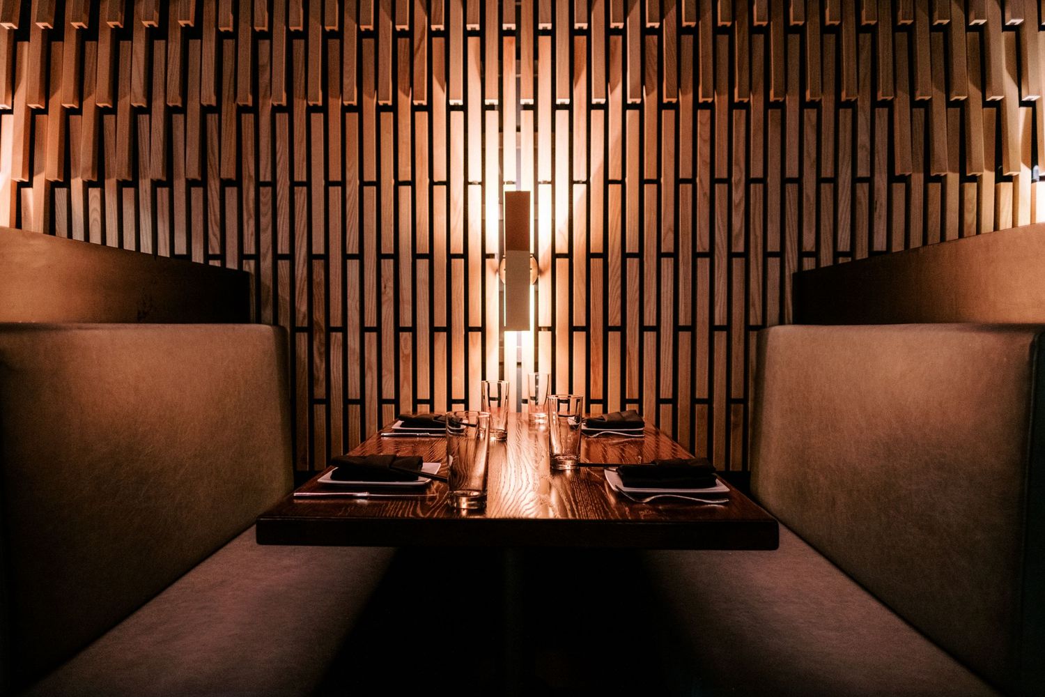 Intimate dining booth with wooden slatted wall and modern table setting under warm lighting.