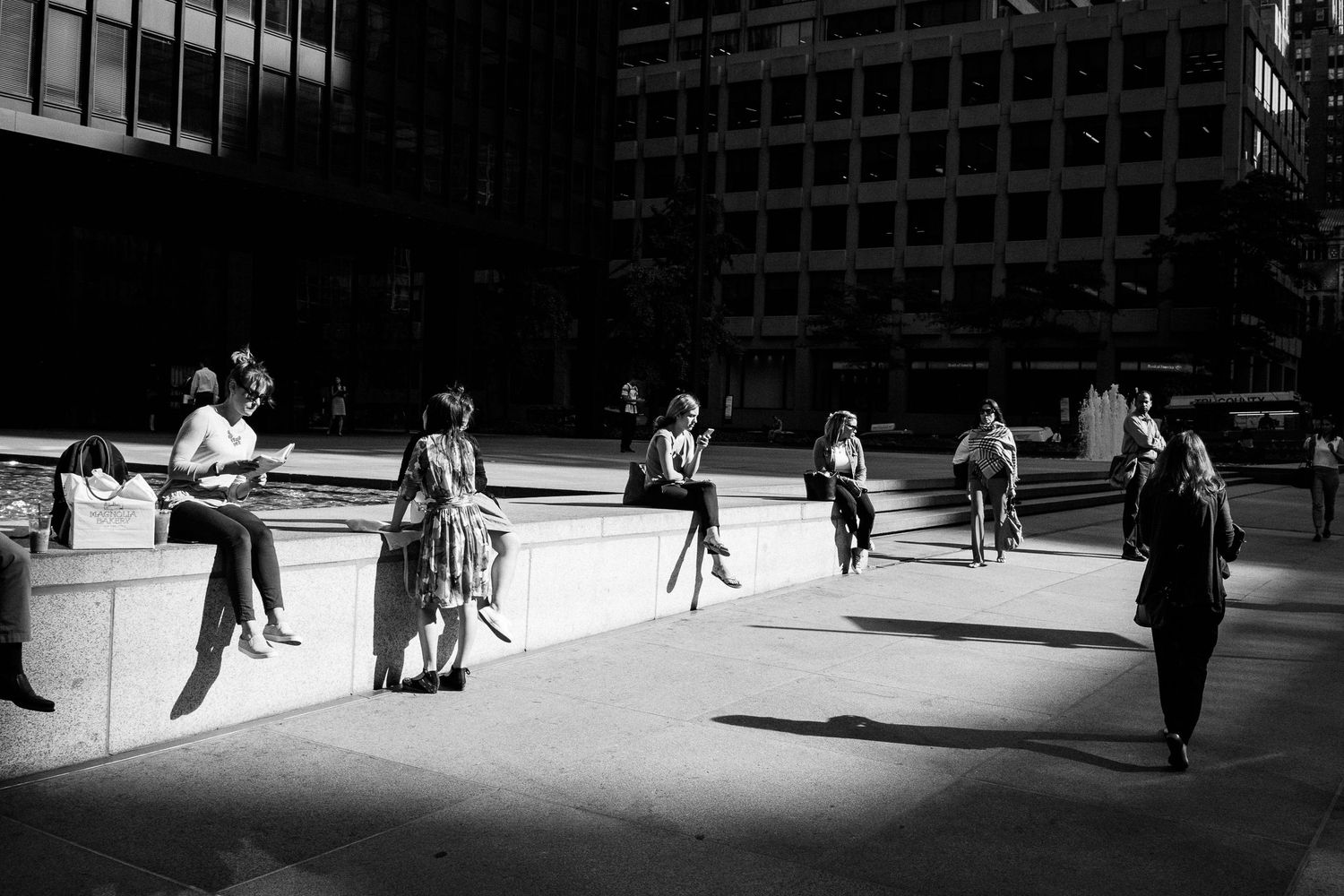 Black and white urban scene showing people sitting and walking in a sunlit plaza with dramatic shadows and modern buildings.