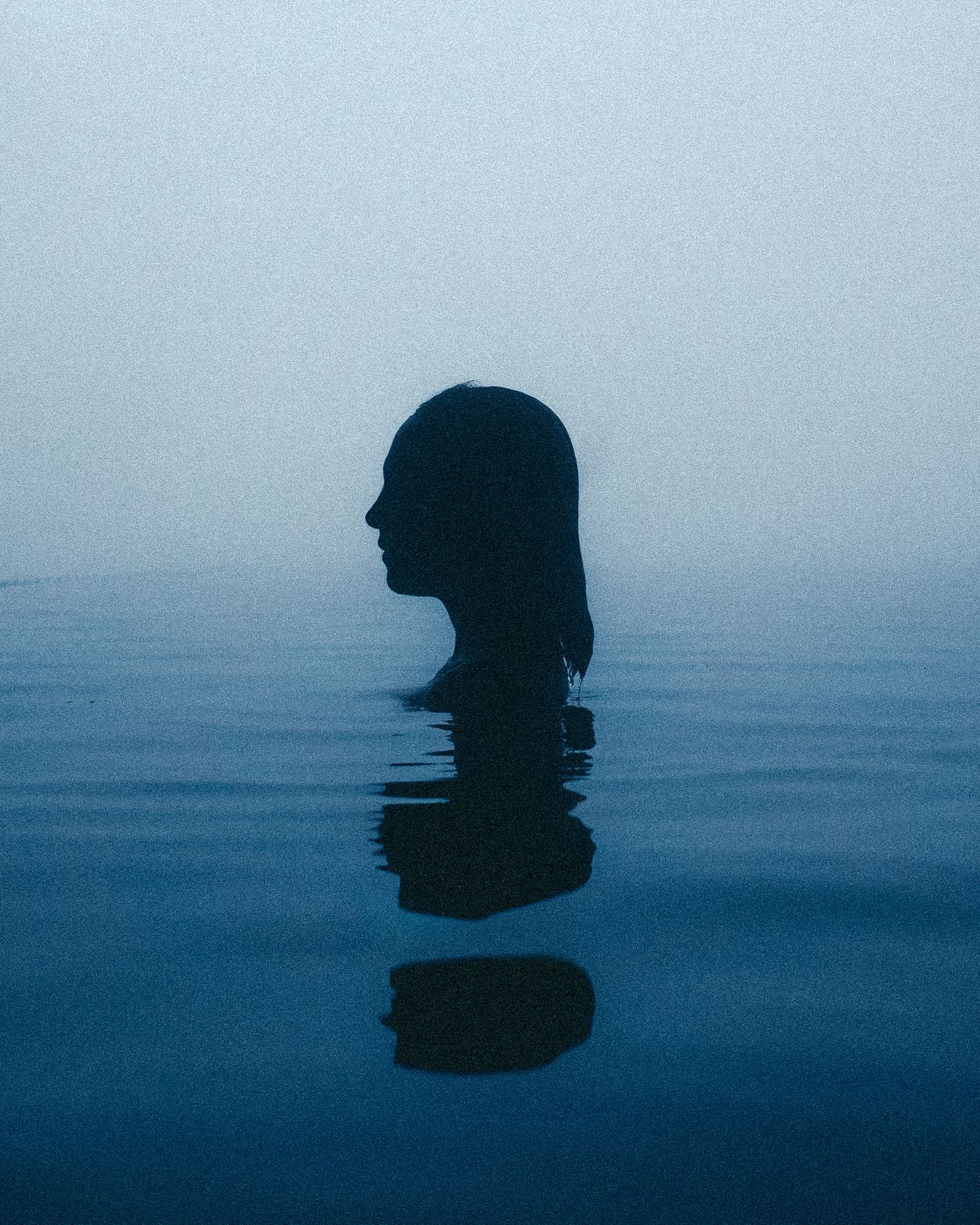 A silhouetted figure in water creates a dark reflection against a misty blue background in a minimalist composition.
