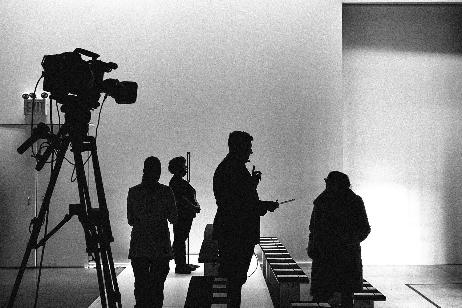 Silhouettes of film crew members and a camera on tripod against bright white background in black and white photo.