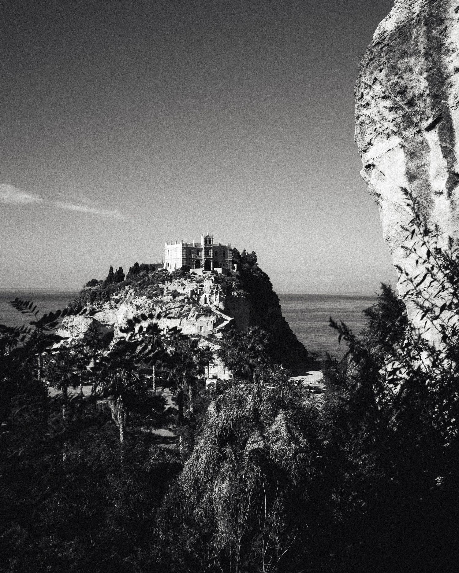 Black and white view of a hilltop castle perched on dramatic coastal cliffs overlooking the Mediterranean Sea.