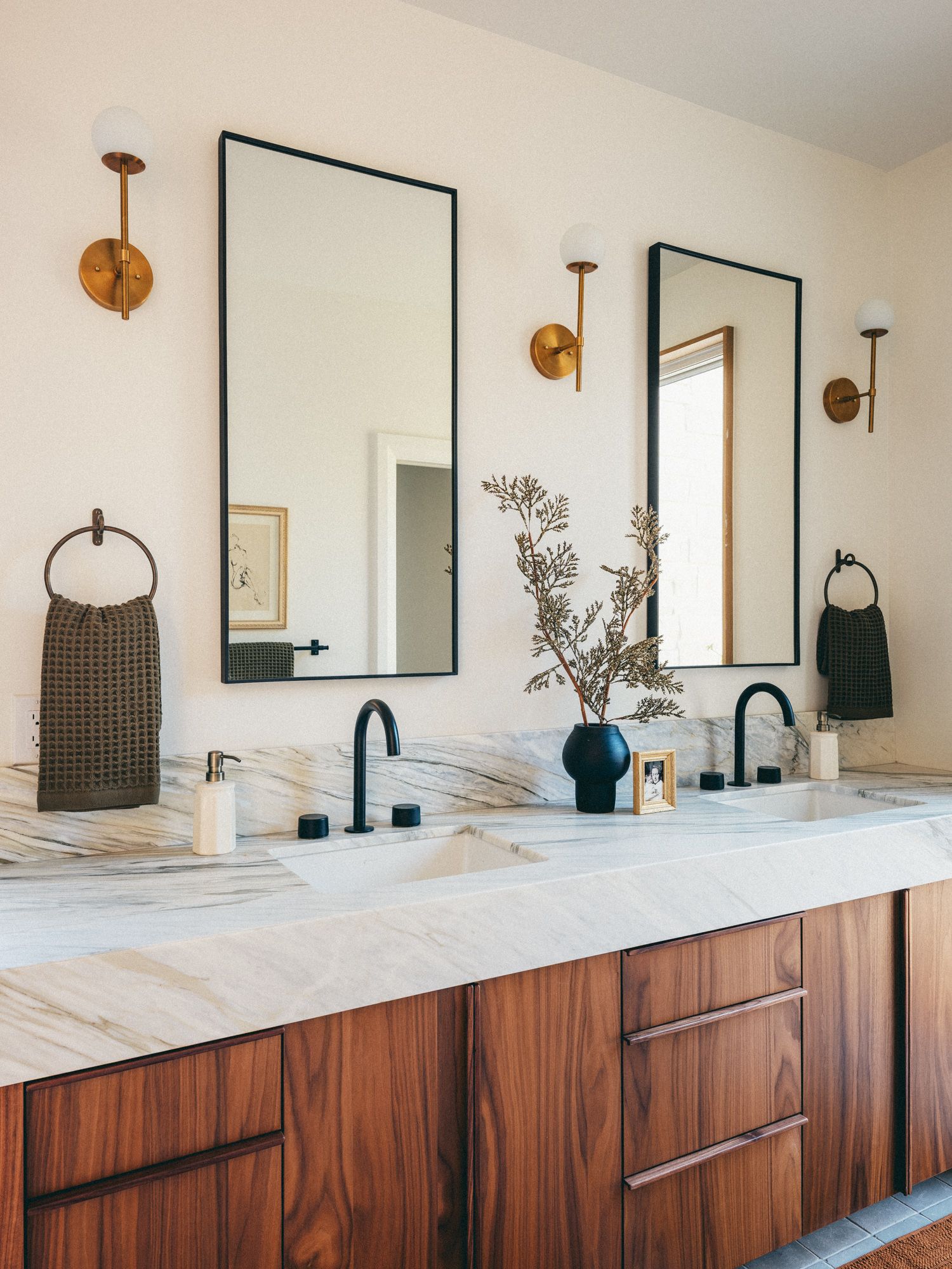 Modern double vanity bathroom with marble countertop, black mirrors, walnut cabinets, and brass wall sconces.