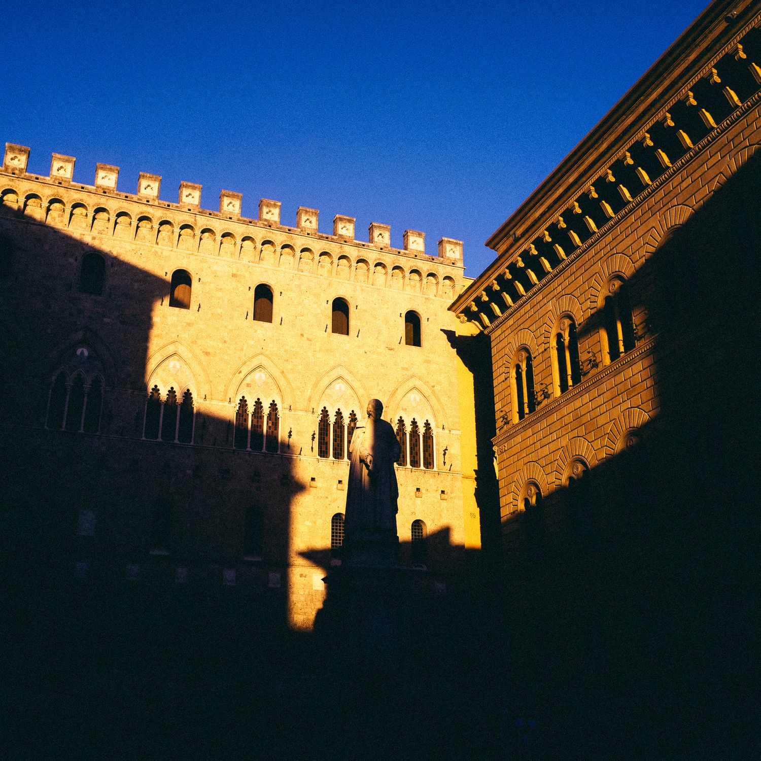 Medieval stone fortress walls and crenellations cast dramatic shadows in golden sunlight against deep blue sky.