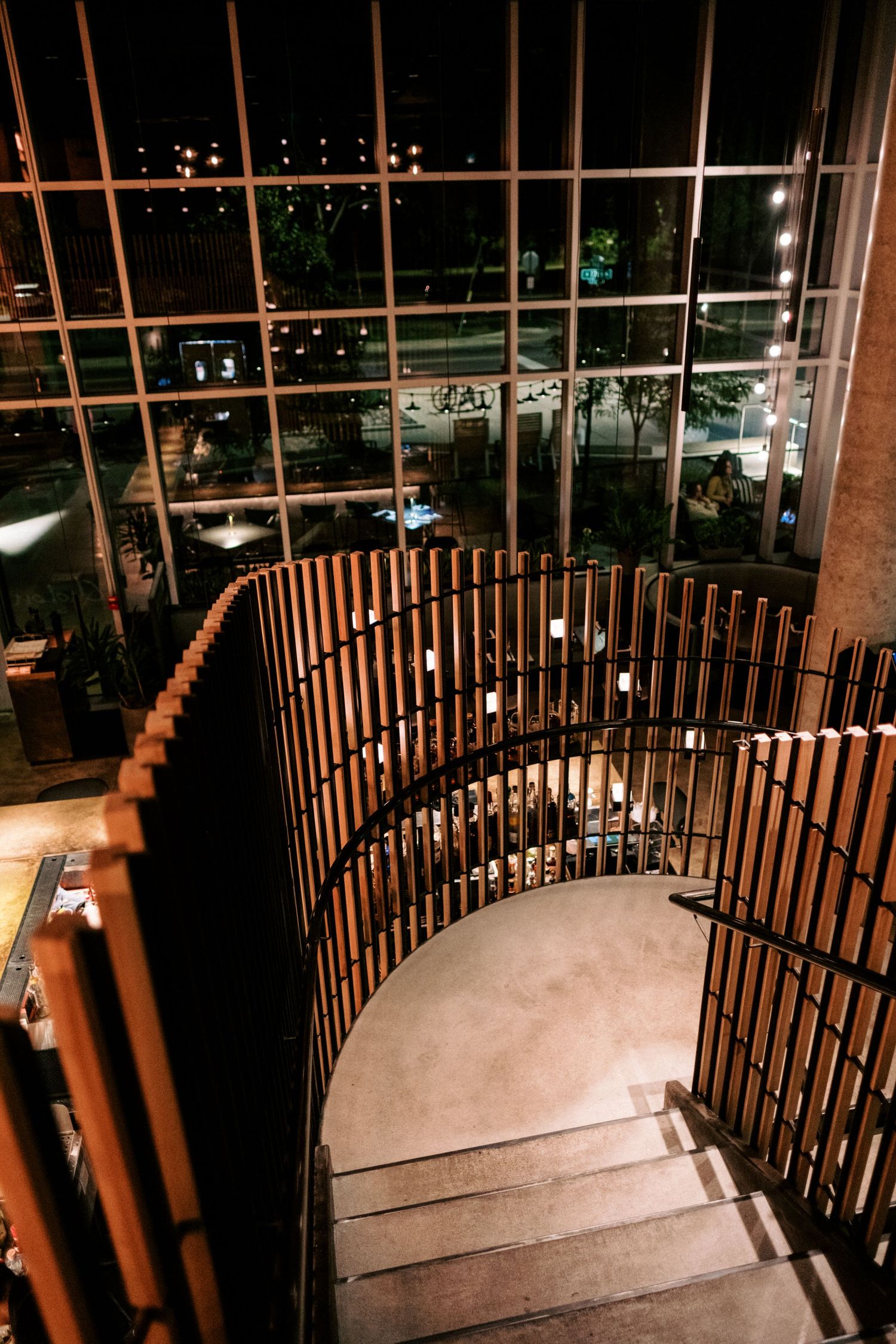 Modern spiral staircase with wooden railings and glass windows overlooking city lights at night.