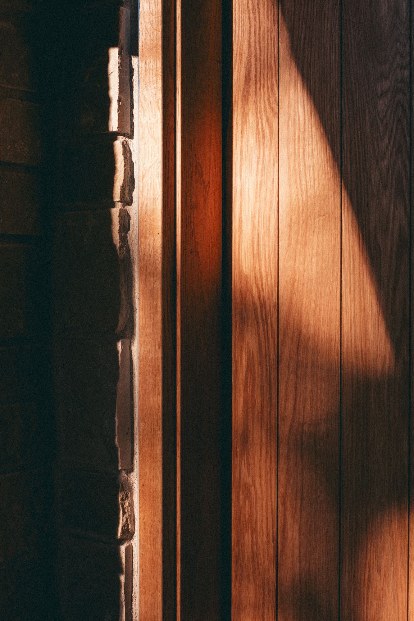 Warm sunlight casts a triangular shadow on a wooden door panel beside a rustic brick wall.