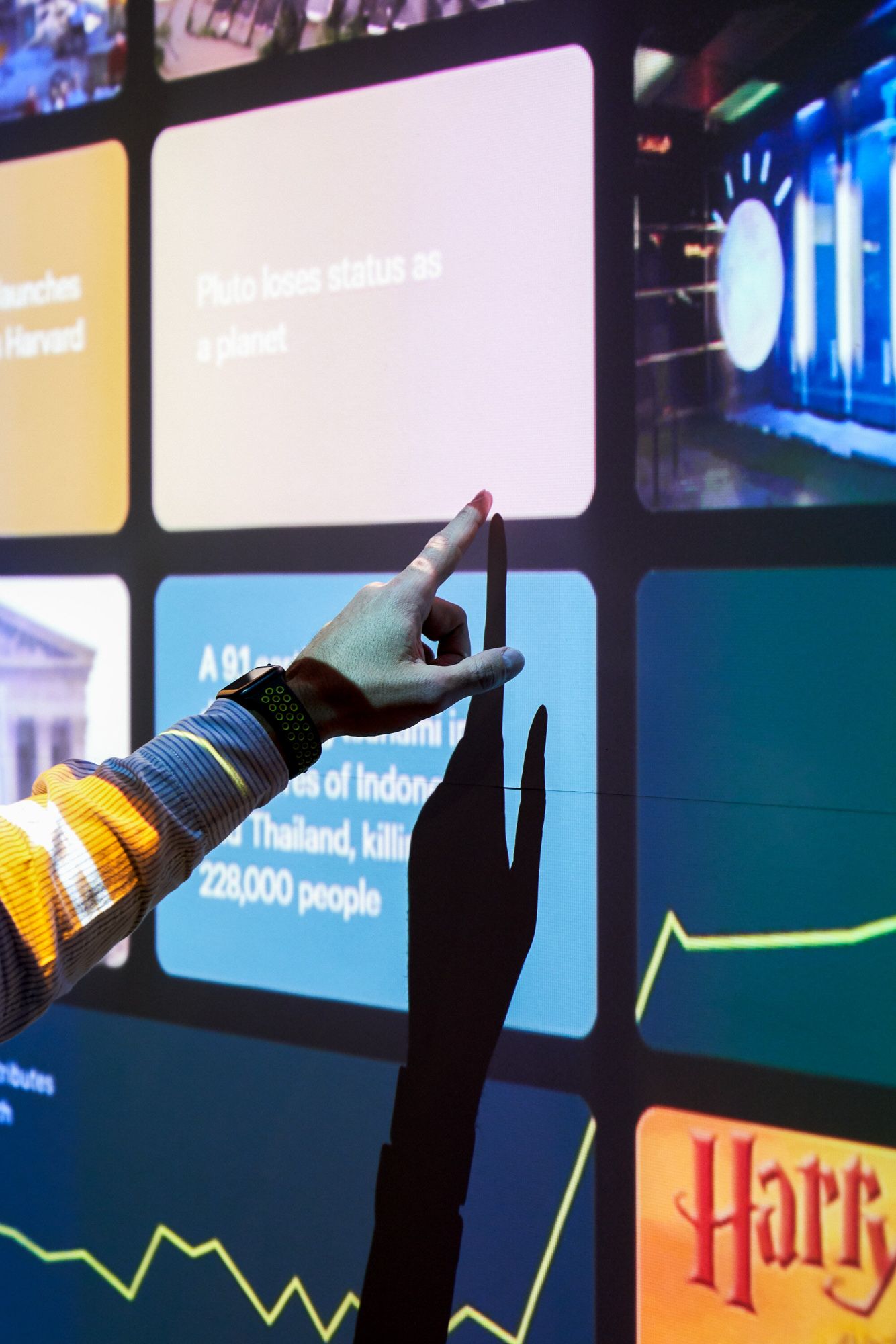 A hand with red nail polish touches an interactive touchscreen display showing news headlines and data charts.