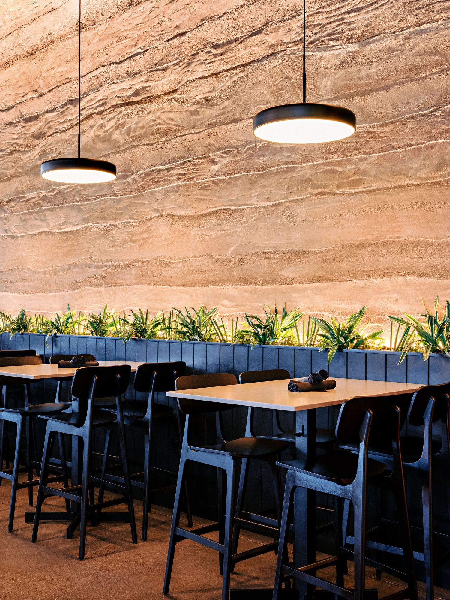 Contemporary dining area with high-top tables and textured stone wall illuminated by pendant lights.