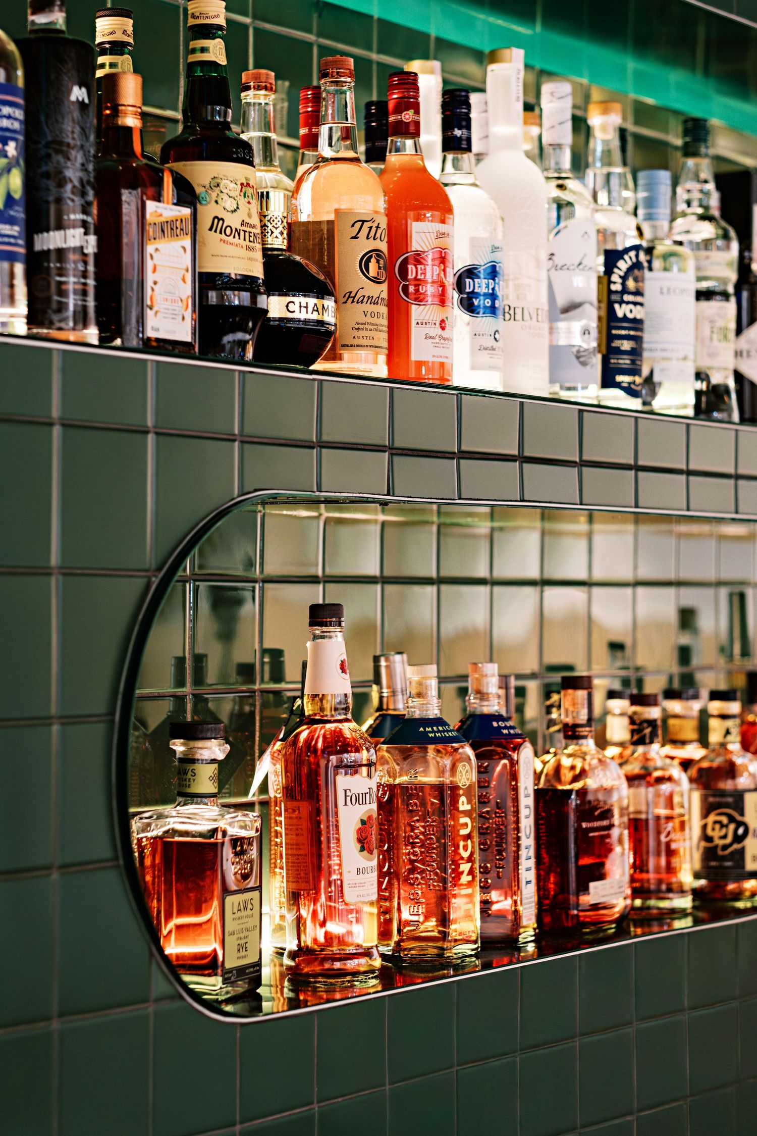 Well-stocked liquor shelf displaying various bottles of spirits with green tile background.