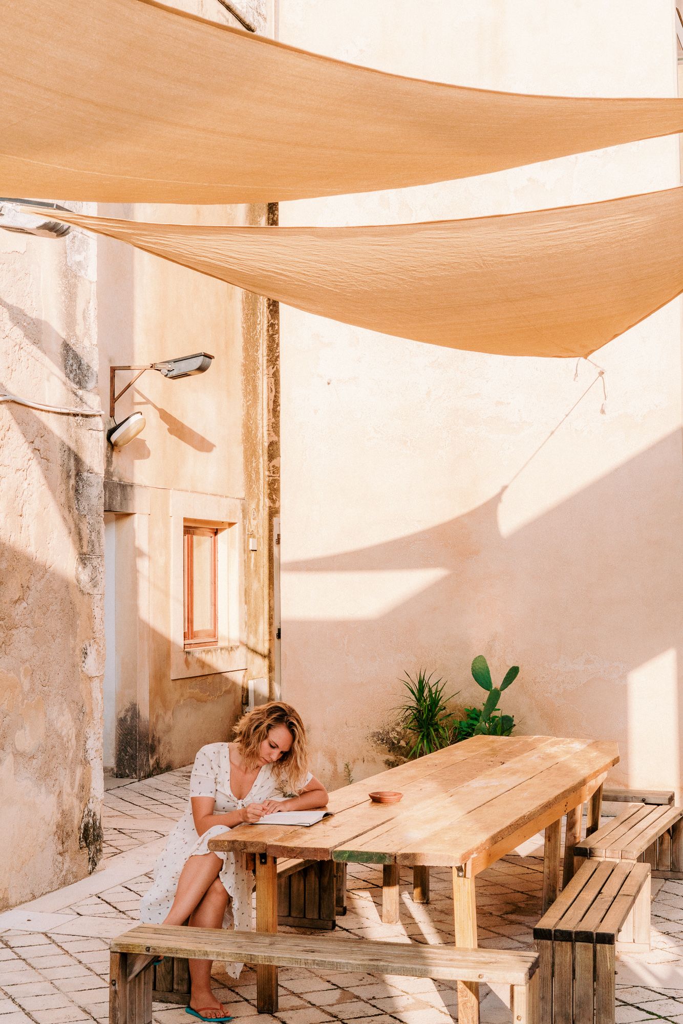 Sun-drenched outdoor patio with wooden benches and beige canvas shade sails.