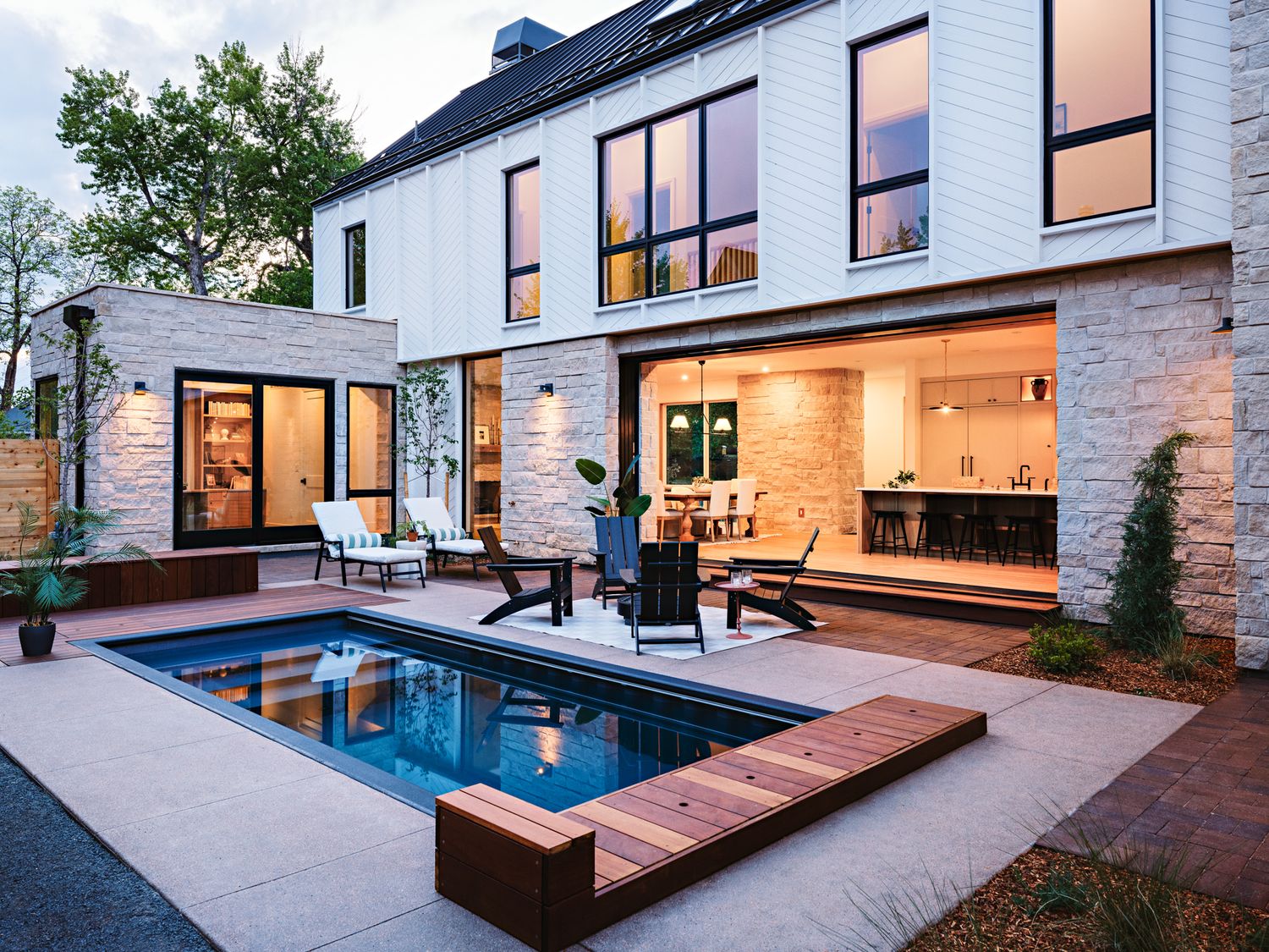 Modern two-story home with small pool, wood deck, outdoor seating, and illuminated open-concept kitchen at dusk.