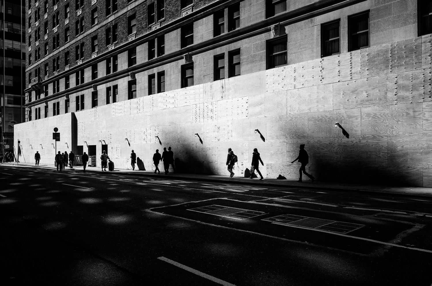 Black and white urban street scene with silhouettes of pedestrians walking against a sunlit wall casting dramatic shadows.