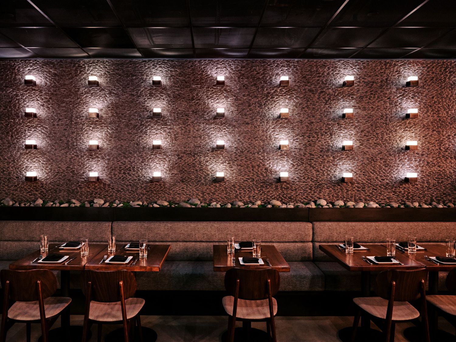 Modern restaurant interior with brick wall featuring grid of warm spotlights and wooden chairs at dining tables.