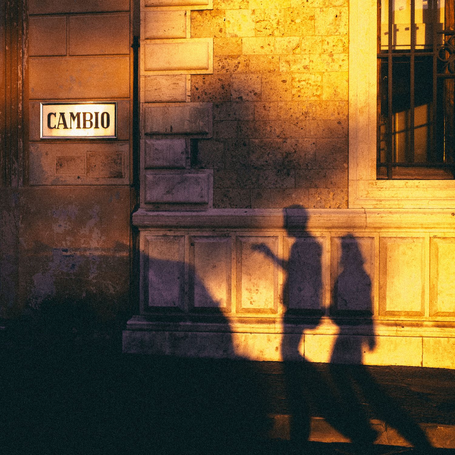 Silhouettes of people cast long shadows against a warmly lit stone building wall at dusk.