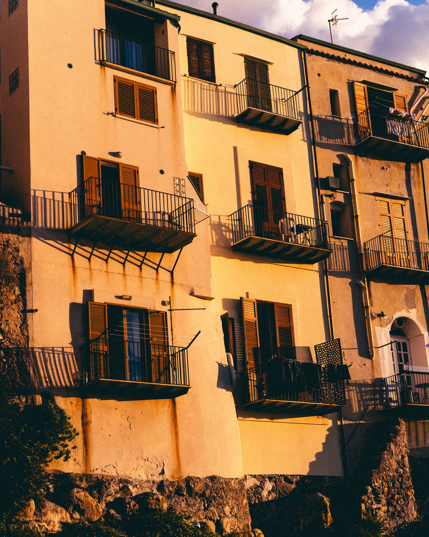 Sunlit Mediterranean-style apartment building with wrought iron balconies and warm yellow walls against a blue sky.