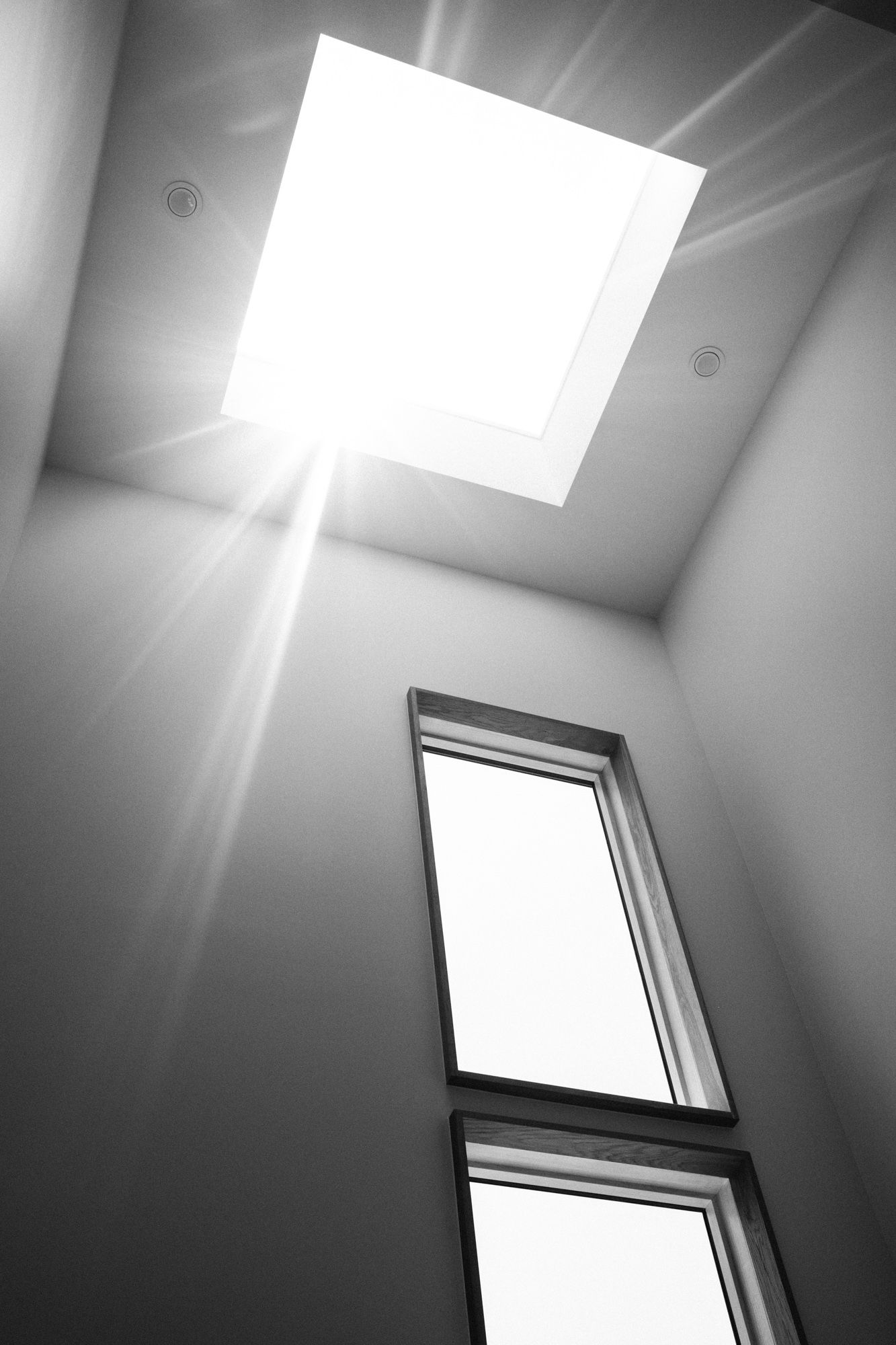 Black and white interior photo looking up at a bright skylight with sun rays and a tall window below.