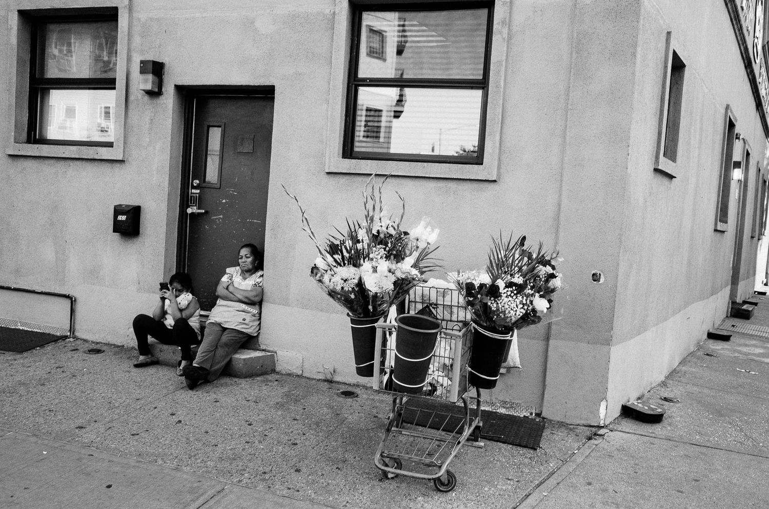 Black and white street scene with shopping carts filled with flowers outside a building with someone sitting by the door.