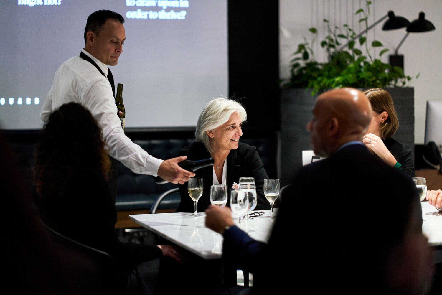 People networking at a modern restaurant meeting with presentation screen and wine glasses on table.