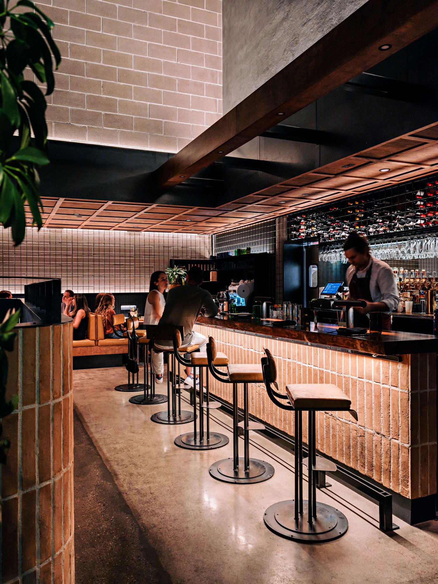 Modern bar counter with sleek bar stools and tiled wall backdrop illuminated by warm lighting.