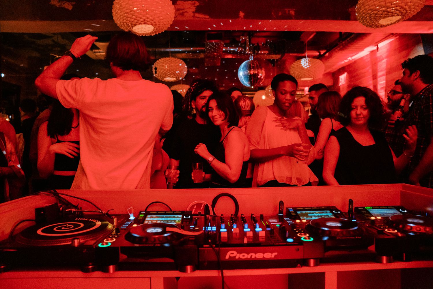 A DJ performs on Pioneer decks for a crowd of dancers in a red-lit nightclub with a disco ball overhead.