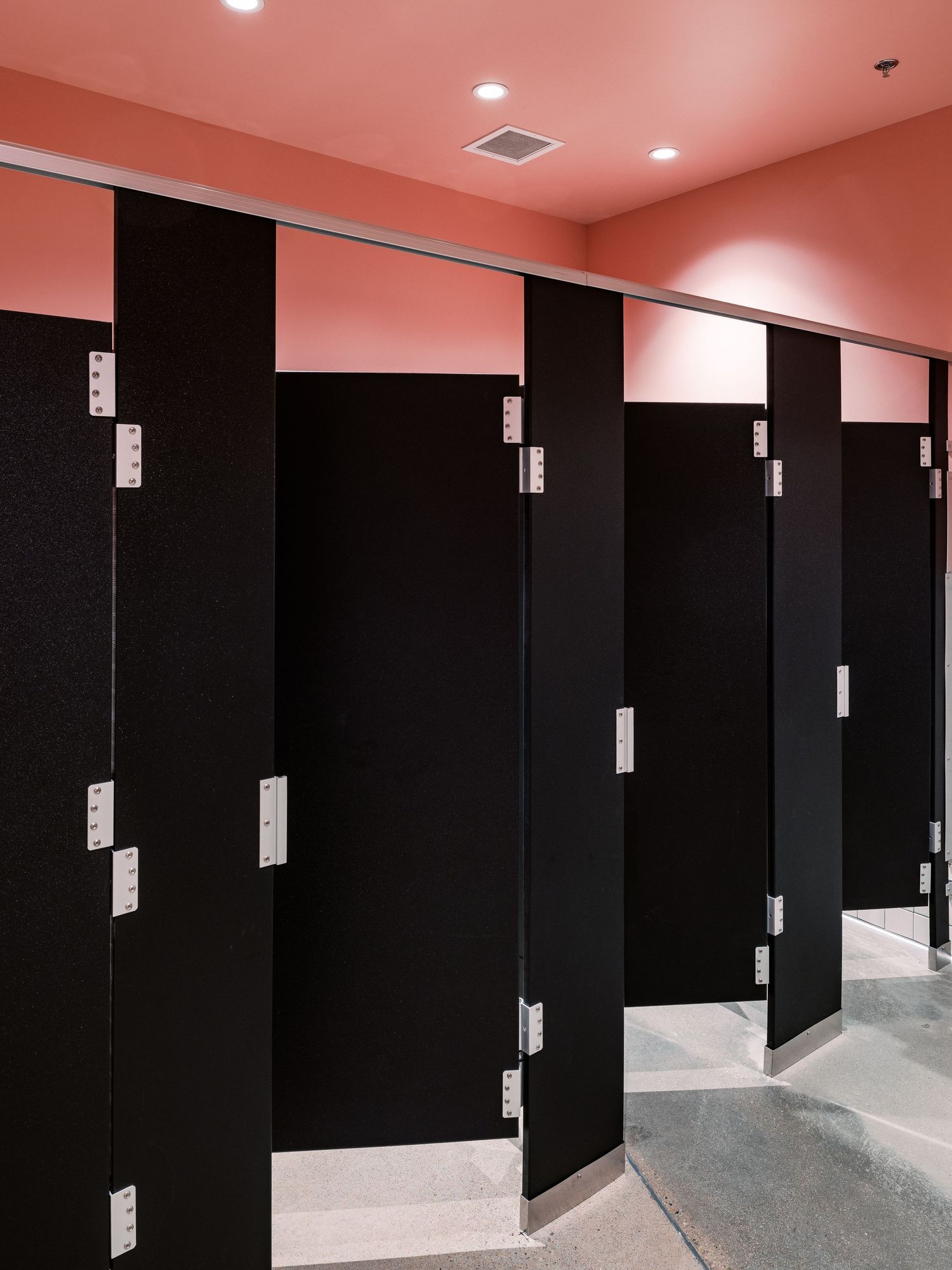 Row of black bathroom stalls with metal hardware in a modern restroom with pink ceiling.