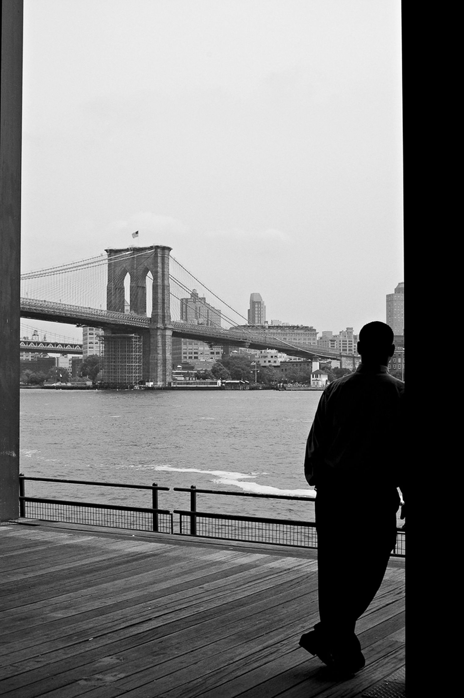 A silhouetted figure gazes at the Brooklyn Bridge from a wooden pier on a misty day.