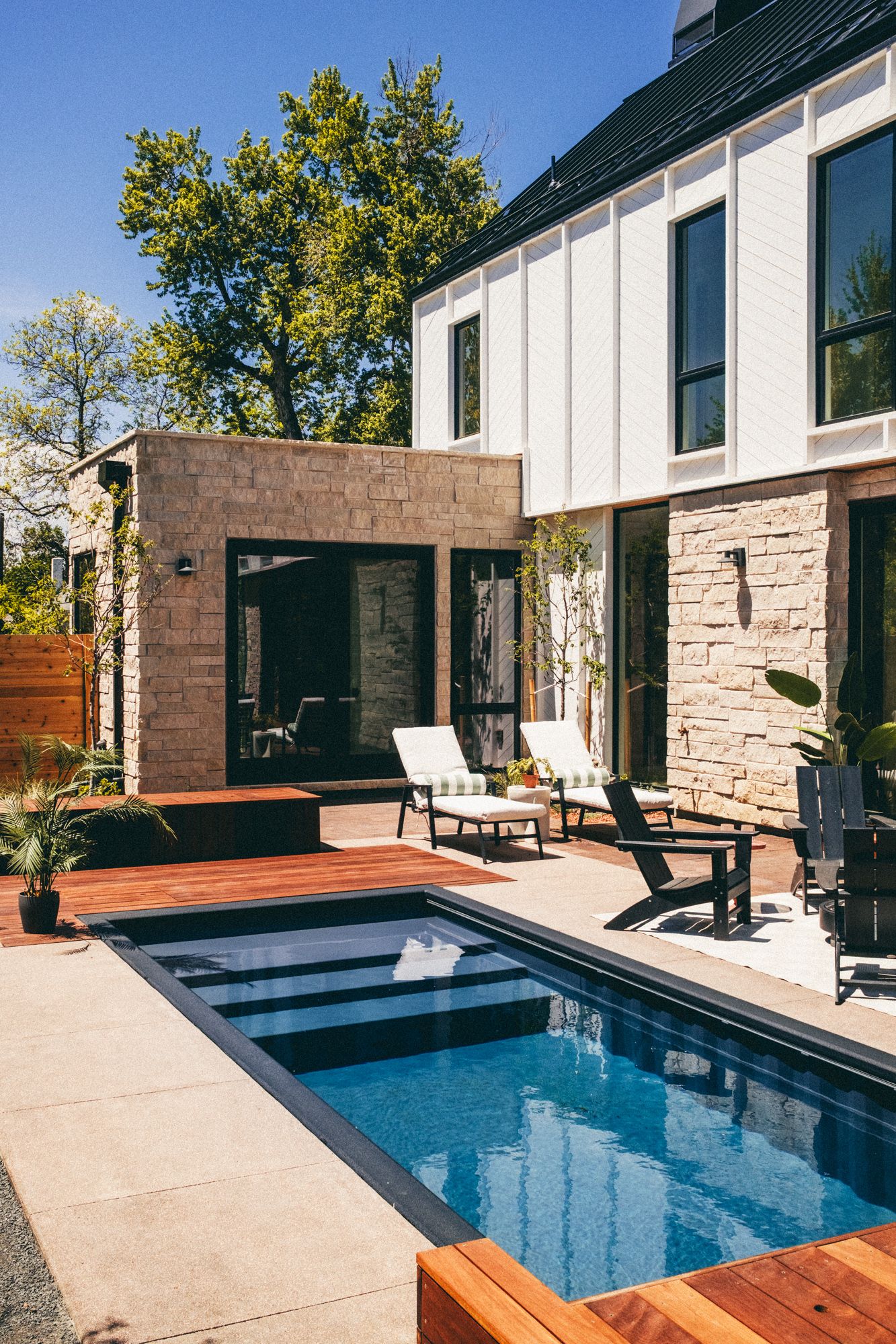 Modern home backyard with small rectangular pool, stone exterior, wood deck, and white lounge chairs on sunny day.