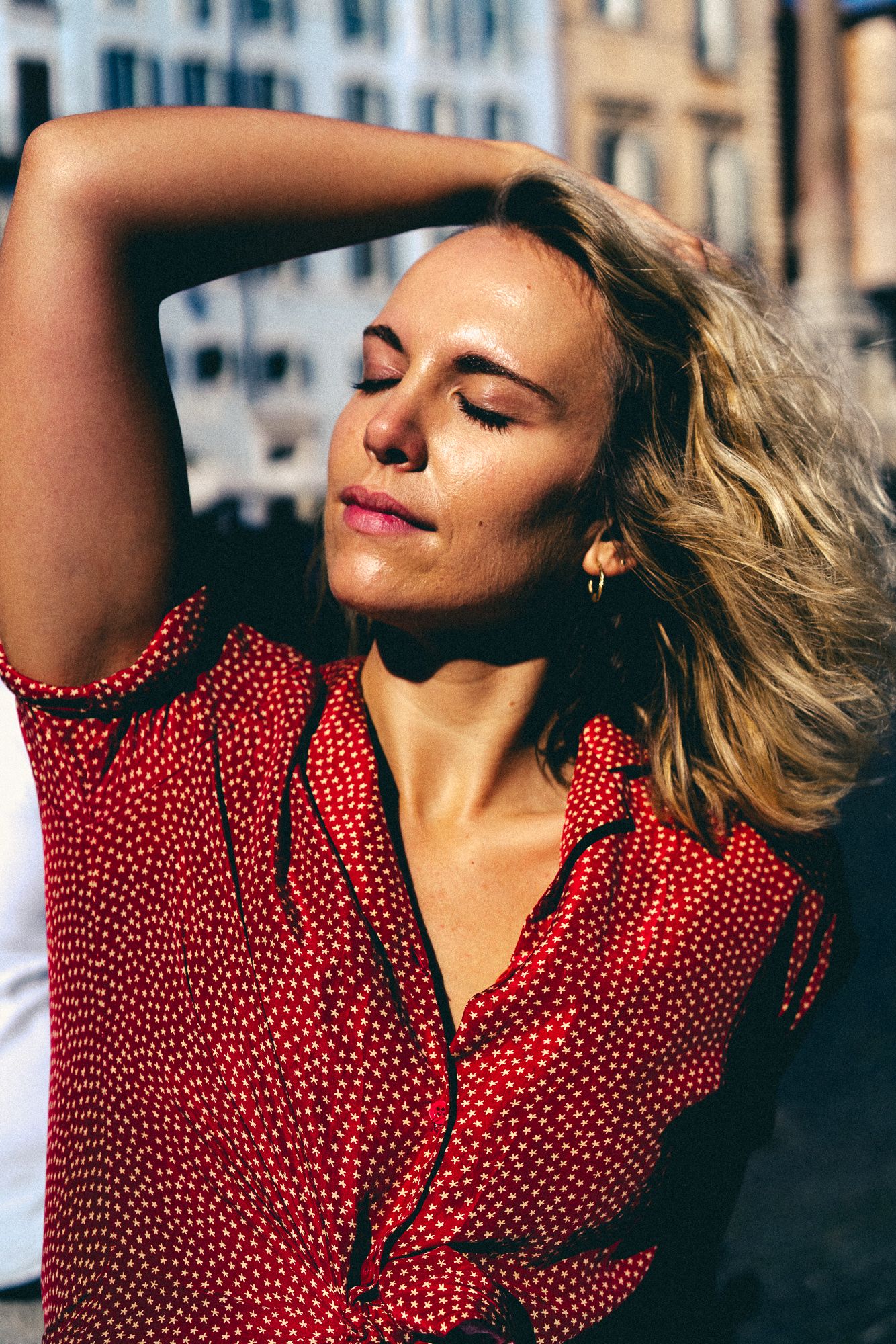 Woman in red polka dot dress enjoying sunlight with hair blowing in the breeze on a city street.