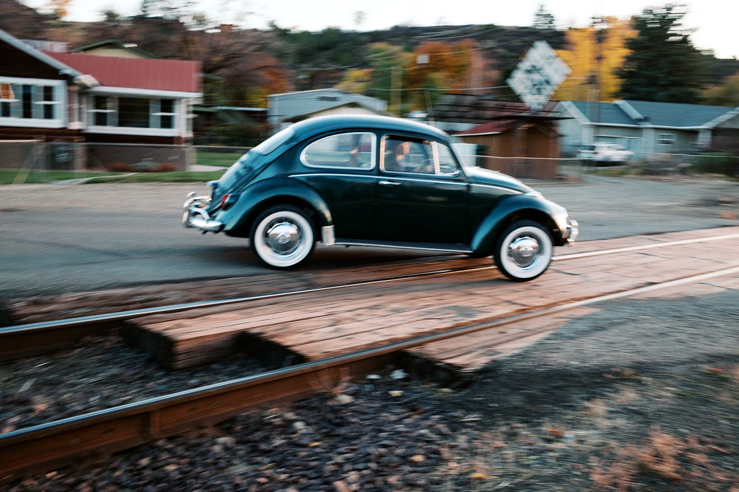 Classic Volkswagen Beetle driving down a street with autumn trees in the background.