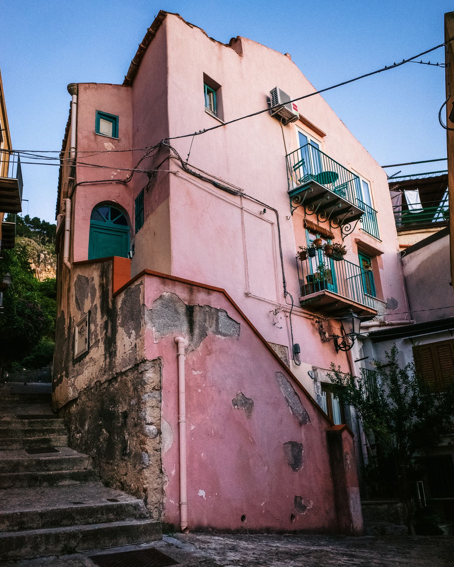 A series of pink-painted buildings with stone stairs and rustic architectural details create a charming Mediterranean streetscape.