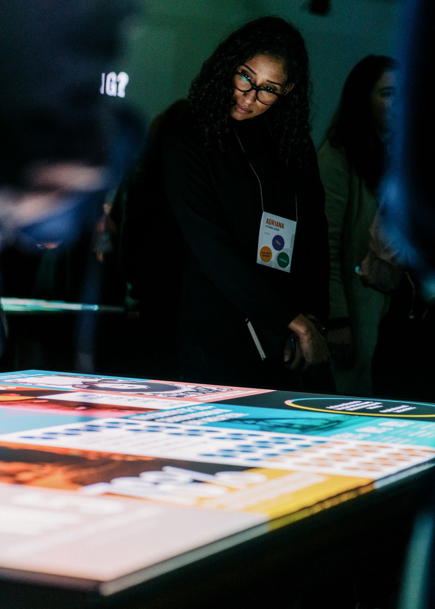 Woman with glasses and lanyard examines an illuminated interactive display table at a tech conference event.