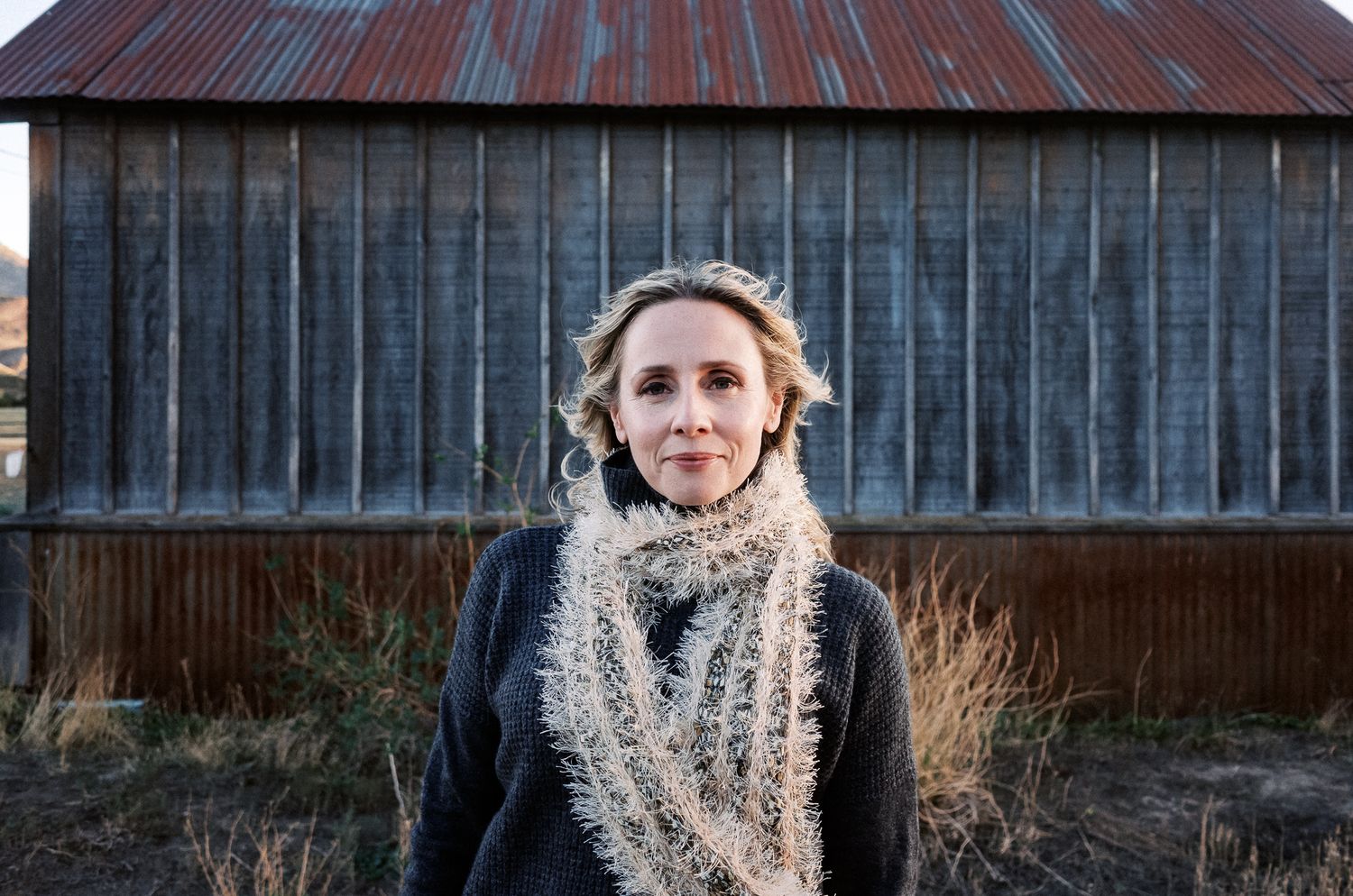 Person wearing winter hat and scarf standing against rustic barn exterior.