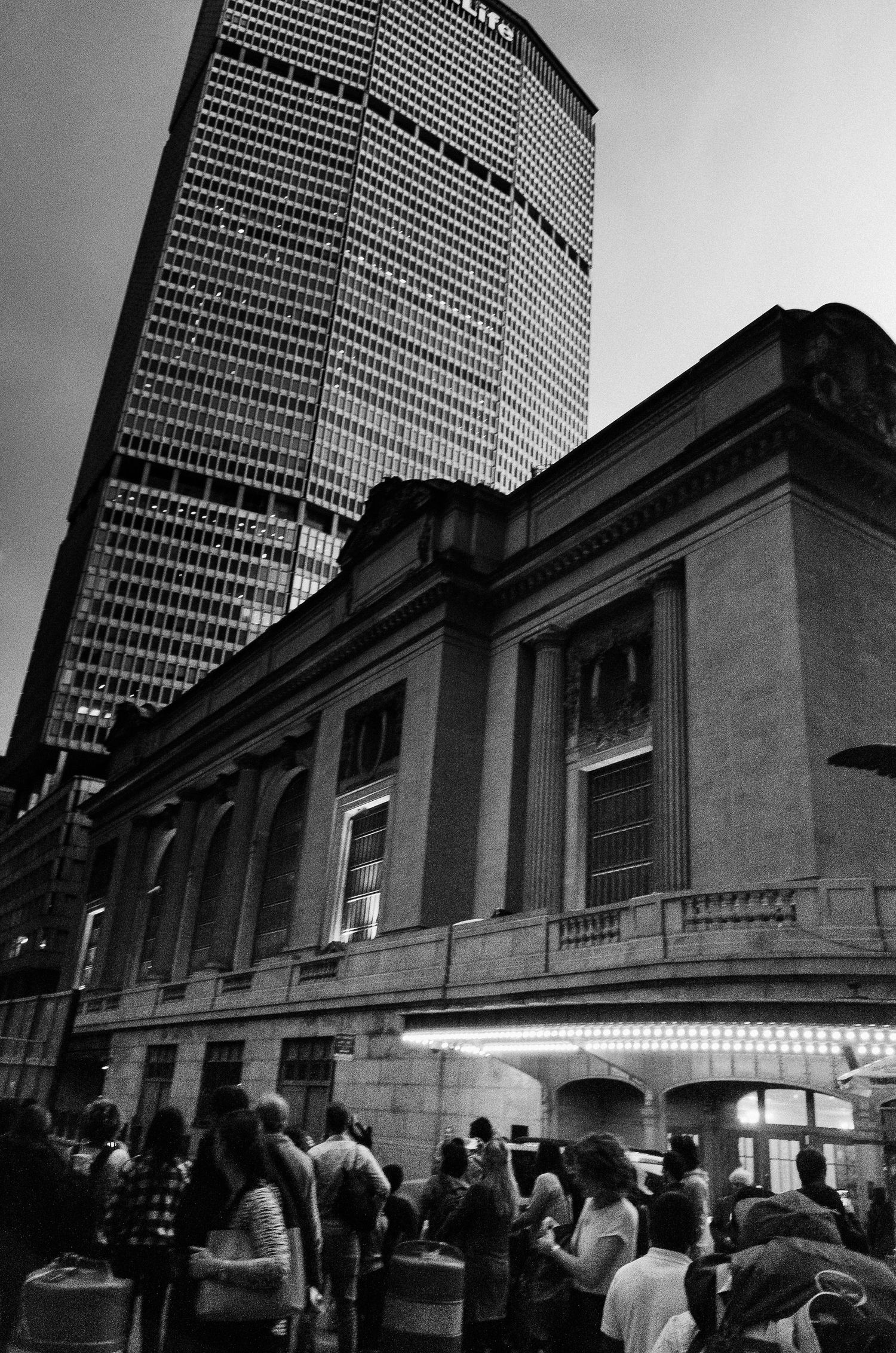 A black and white photo of the MetLife Building towering over Grand Central Terminal with crowds gathering below.